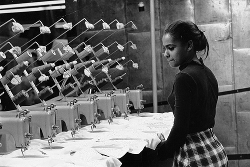 A young African American woman watches at least seven sewing machines working simultaneously, de Kheel Center